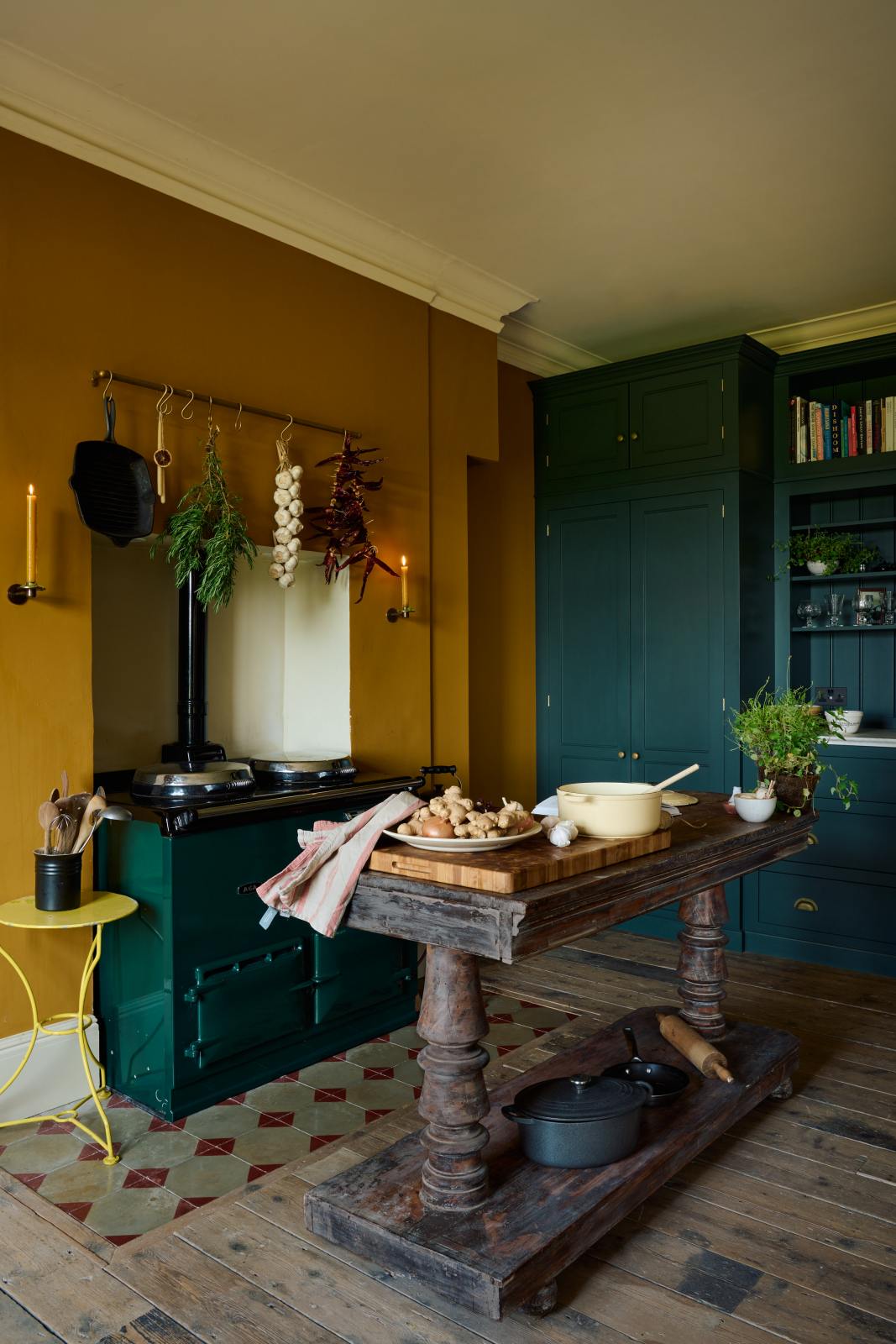 An angled view of a lovely modern kitchen that retains the charm of its original structure and flooring, using primary colours in a muted, mature palette. The spacious kitchen features a Clerkenwell Blue full-height pantry cabinet and a long sink run with a marble worktop. An emerald green gas stove sits within the chimney breast, accompanied by a deVOL cream pendant light above a vintage round wooden table, with matching chairs and a prep table.