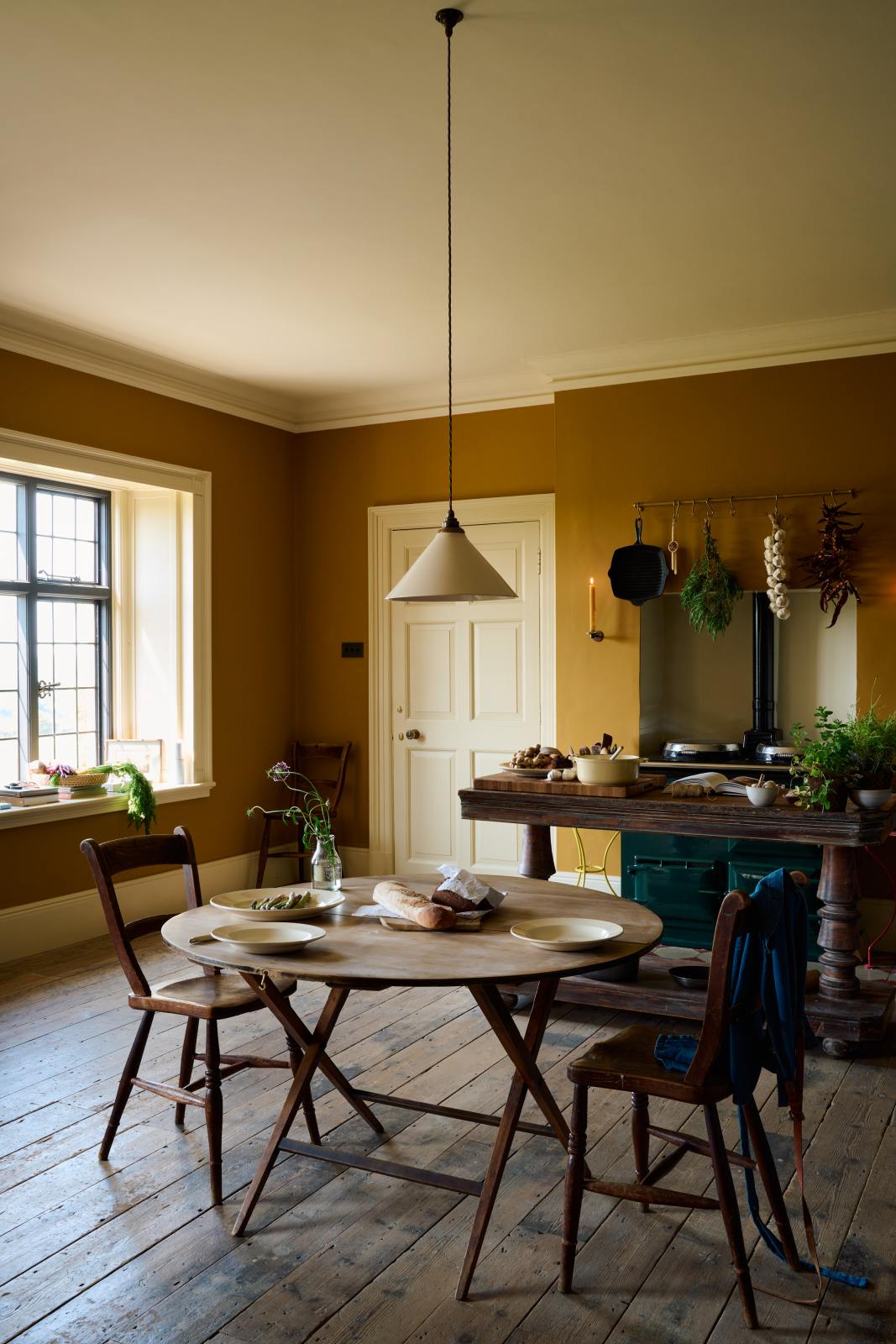 A closer look at a lovely modern kitchen that retains the charm of its original structure and flooring, using the three primary colours in a muted, mature palette. The spacious kitchen features an emerald green gas stove sits within the chimney breast, accompanied by a deVOL cream pendant light above a vintage round wooden table, with matching chairs and a prep table.