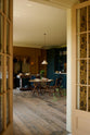 Looking through the french doors is a lovely modern kitchen that retains the charm of its original structure and flooring, using the three primary colours in a muted, mature palette. The spacious kitchen features a Clerkenwell Blue full-height pantry cabinet, a long sink run with a marble worktop, and an open countertop cabinet and shelf. A deVOL cream pendant light above a vintage round wooden table, with matching chairs and a vintage prep table.