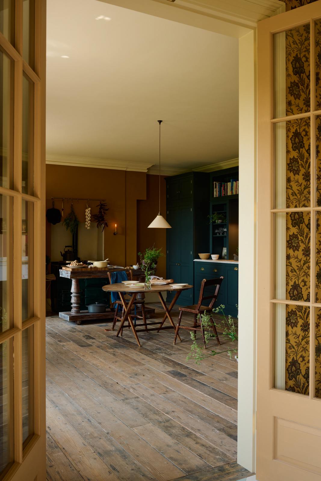 Looking through the french doors is a lovely modern kitchen that retains the charm of its original structure and flooring, using the three primary colours in a muted, mature palette. The spacious kitchen features a Clerkenwell Blue full-height pantry cabinet, a long sink run with a marble worktop, and an open countertop cabinet and shelf. A deVOL cream pendant light above a vintage round wooden table, with matching chairs and a vintage prep table.