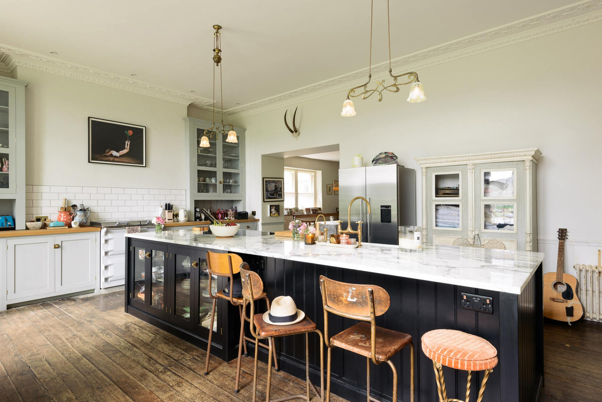 A tall-ceilinged classic English kitchen centres around a wide Printer’s Black island with an aged brass Ionian tap, marble worktop, and glazed base cabinets. A cosy breakfast nook with vintage stools sits opposite an off-white cooker run and matching wall cabinets. The space feels artfully lived-in—funky paintings, a guitar, and kitchen essentials tell the story of a creative, full-of-life family.