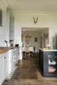 The tall-ceilinged, classic English kitchen centres around a wide painter’s black island with a marble worktop, glazed base cabinets, an off-white cooker run, and matching wall cabinets. An archway leads to a large dining room with a vintage lace tablecloth on a wooden dining table, vintage chairs, and a square lace chandelier with tassels. The space feels artfully lived-in—funky paintings, hanging sculptures, and kitchen essentials tell the story of a creative, full-of-life family.