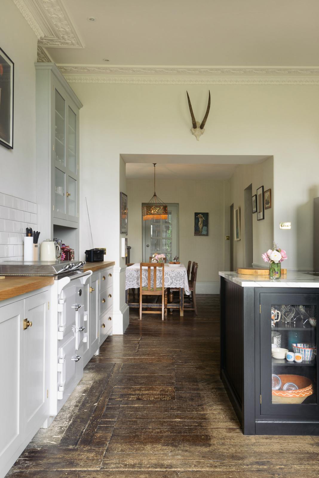 The tall-ceilinged, classic English kitchen centres around a wide painter’s black island with a marble worktop, glazed base cabinets, an off-white cooker run, and matching wall cabinets. An archway leads to a large dining room with a vintage lace tablecloth on a wooden dining table, vintage chairs, and a square lace chandelier with tassels. The space feels artfully lived-in—funky paintings, hanging sculptures, and kitchen essentials tell the story of a creative, full-of-life family.