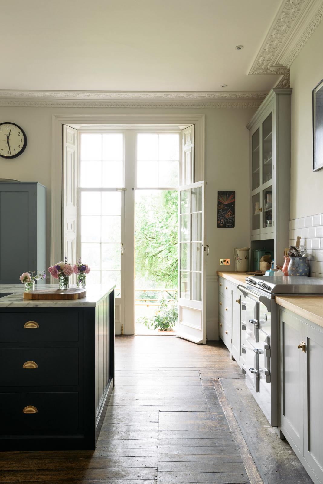 One side of a French door is left open to the lush green garden, allowing bright white natural light to pour into the tall-ceilinged, classic English kitchen centred around a wide painter’s black island with a marble worktop, and an off-white cooker run with matching wall cabinets and white porcelain splashback tiles.