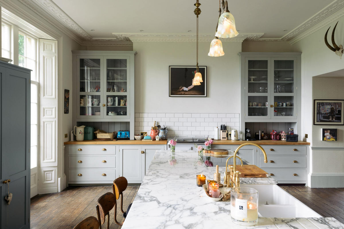 A tall-ceilinged, classic English kitchen features a wide painter’s black island with an aged brass Ionian tap, marble worktop, a cosy breakfast nook with vintage stools, an off-white cooker run, and matching wall cabinets. The space feels artfully lived-in — funky paintings, hanging sculptures, delicate glass gaselier light, and kitchen essentials tell the story of a creative, lively family.