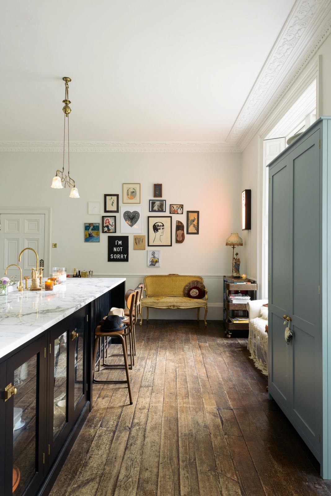 A tall-ceilinged classic English kitchen centres around a painter’s black island with a marble worktop, aged brass Ionian tap, and a full-height pantry cabinet. A cosy breakfast nook includes vintage stools, a gold-framed mustard sofa, and a white one by the window, draped in lace. Funky art, hanging sculptures, a vintage book trolley, and a delicate glass gaselier light give the space a creative, lived-in feel.