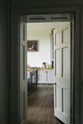 The doorway to a tall-ceilinged, classic English kitchen is dark and nearly open. At the centre of the kitchen is a wide black island with a marble countertop, surrounded by white porcelain splashback tiles, an off-white range cooker, and matching wall cabinets. Above the gas cooker hangs an unusual painting depicting a person being lifted by their legs by a red and green balloon.