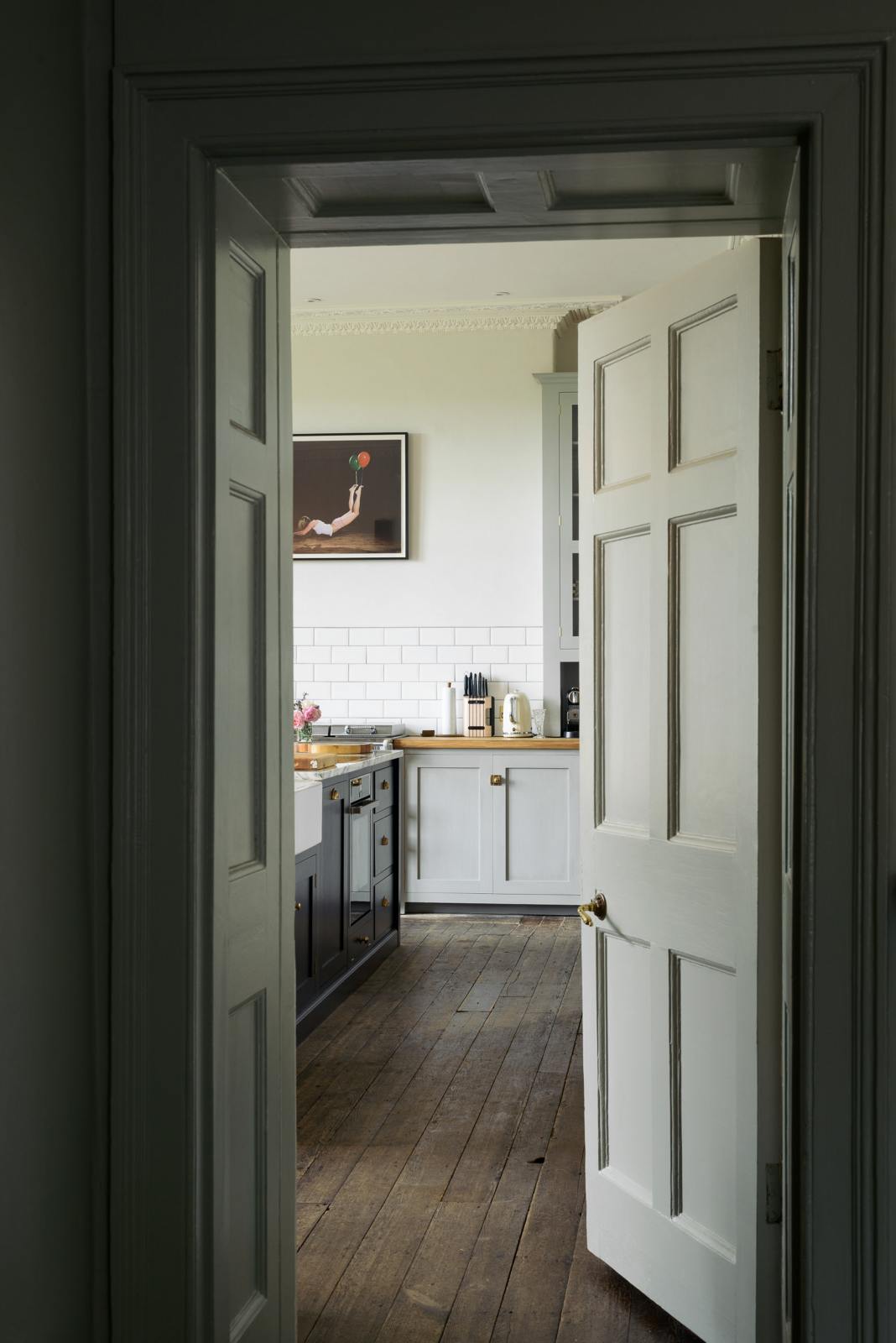 The doorway to a tall-ceilinged, classic English kitchen is dark and nearly open. At the centre of the kitchen is a wide black island with a marble countertop, surrounded by white porcelain splashback tiles, an off-white range cooker, and matching wall cabinets. Above the gas cooker hangs an unusual painting depicting a person being lifted by their legs by a red and green balloon.