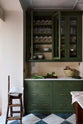 A closer look at a spacious and elegant Wilkes green classic English kitchen, featuring a long L-shaped run with a cooker and sink, complemented by a Silestone worktop and glazed wall cupboards. Adorned with classic marble chequered-board flooring, cream subway splashback tiles, artisan ceramics, vintage furniture, and luscious green vegetables, it embodies a classic and stylish aesthetic.