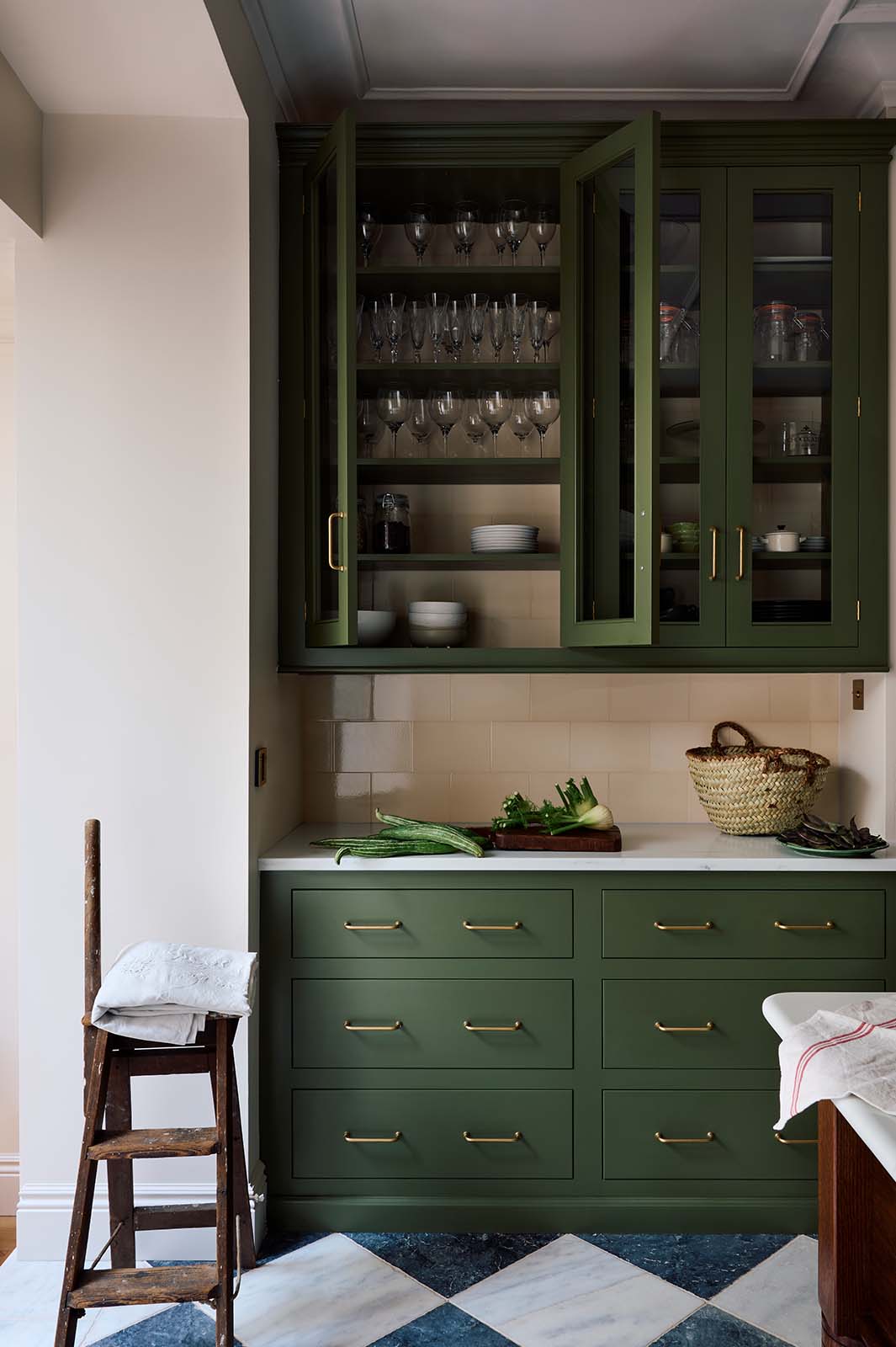 A closer look at a spacious and elegant Wilkes green classic English kitchen, featuring a long L-shaped run with a cooker and sink, complemented by a Silestone worktop and glazed wall cupboards. Adorned with classic marble chequered-board flooring, cream subway splashback tiles, artisan ceramics, vintage furniture, and luscious green vegetables, it embodies a classic and stylish aesthetic.