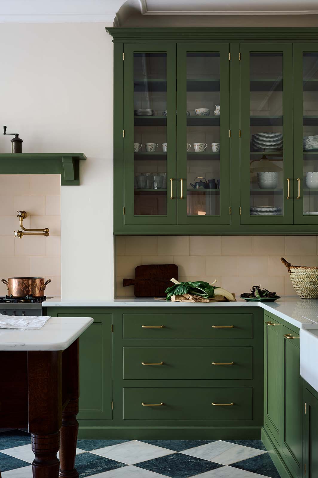 A spacious and elegant Wilkes green classic English kitchen features a long L-shaped cooker and sink run with a Silestone worktop, glazed wall cupboards, shelves, and an Heirloom dairy table. Adorned with classic marble chequered-board flooring, Heirloom task lights, cream subway splashback tiles, copper pots, and luscious green vegetables, it creates a classic and stylish kitchen.