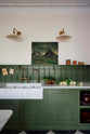 A closer look at a spacious and elegant Wilkes green classic English kitchen featuring a sink run with a double Belfast sink, an aged brass Ionian tap, a low tongue-and-groove panel, and a long shelf with hanging rail. Adorned with vintage containers, Heirloom task lights, a moody landscape painting, and luscious green vegetables, it creates a classic and stylish kitchen.