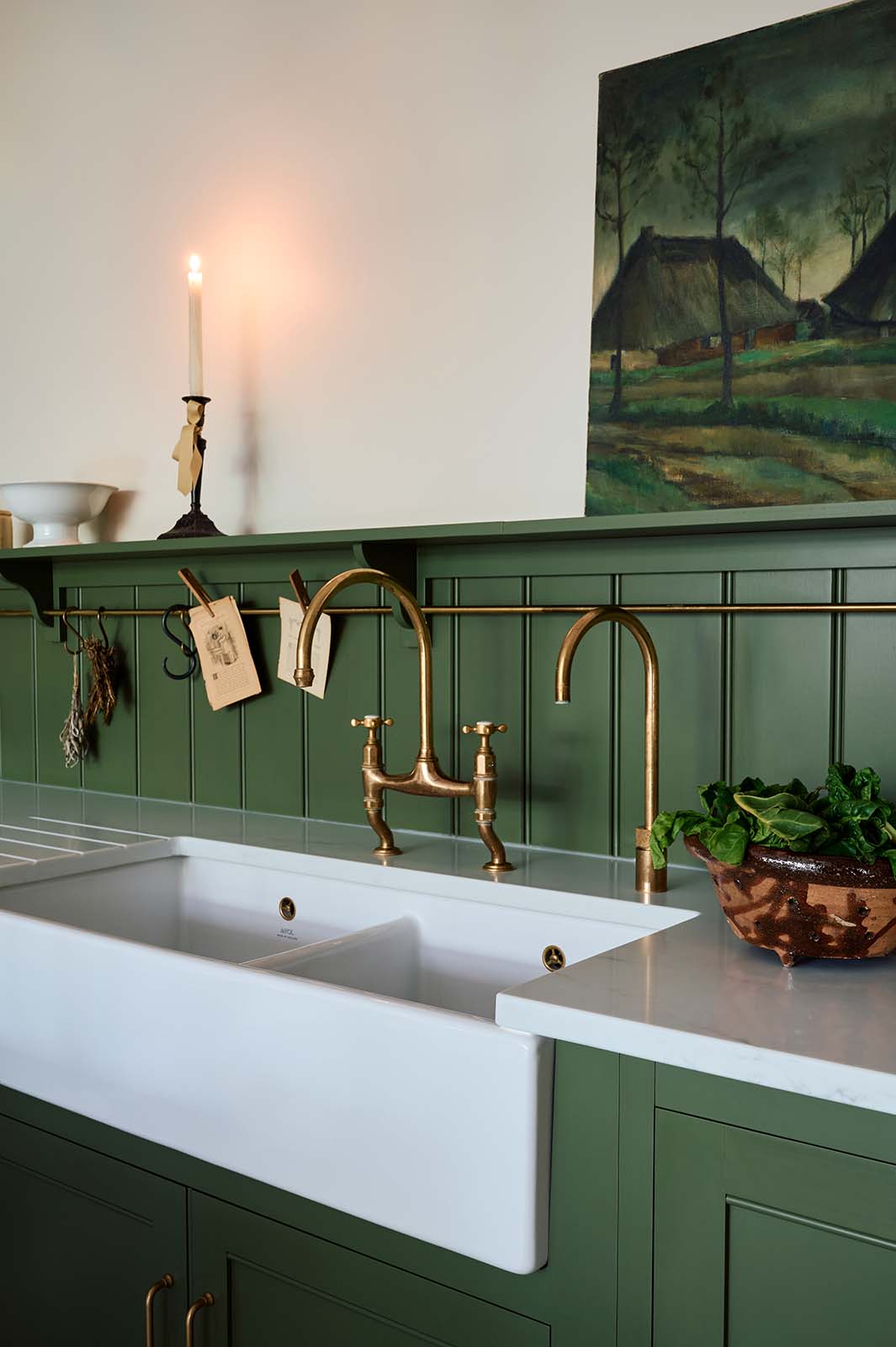 An oblique view of a spacious and elegant Wilkes green classic English kitchen featuring a sink run with a double Belfast sink, an aged brass Ionian tap, a low tongue-and-groove panel, and a long shelf with hanging rail. Adorned with vintage ceramics, Heirloom task lights, a moody landscape painting, and luscious green vegetables, it creates a classic and stylish kitchen.