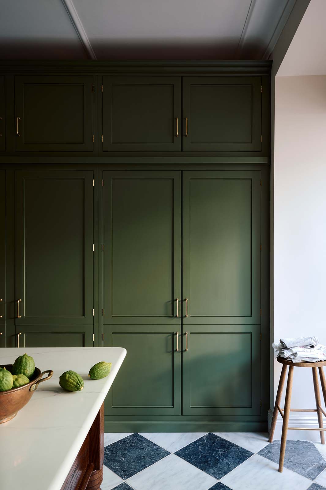 A spacious and elegant Wilkes green classic English kitchen features a bank of full-height pantry cupboards, and an Heirloom dairy table. Adorned with classic marble chequered-board flooring, a bowl of limes, a Helen stool, and tea towels, it creates a classic and stylish kitchen.