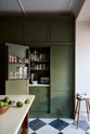A spacious and elegant Wilkes green classic English kitchen features a bank of full-height pantry cupboards, and an Heirloom dairy table. Adorned with classic marble chequered-board flooring, a bowl of limes, a Helen stool, dreid ingredients, glass containers, and tea towels, it creates a classic and stylish kitchen.