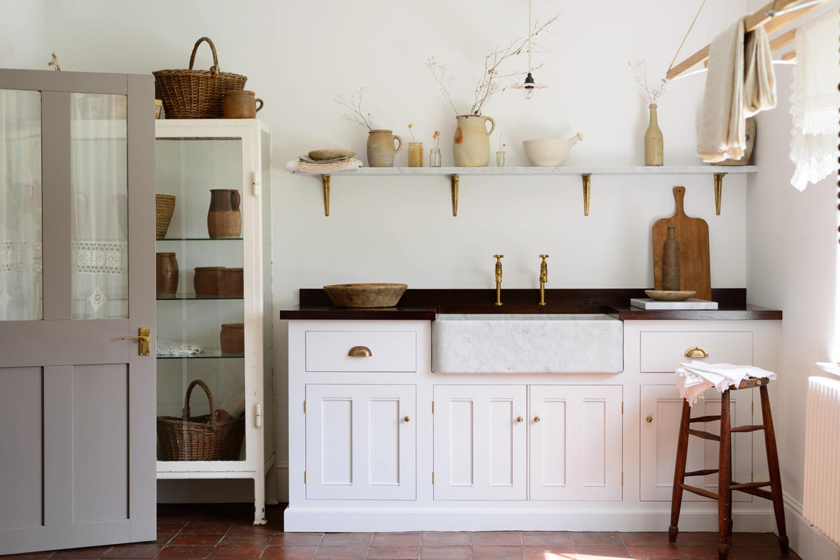 The simple and functional linen white scullery room with a modest sink run with polished sapele, a large single Carrara marble sink, and aged brass Mayan taps. The warm reds and browns of the terracotta square tiles, vintage furniture, ceramics, woven baskets and a deVOL laundry maid serve the room with both a purpose and beautiful aesthetics.