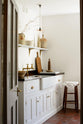 An angled view of a simple and functional linen white scullery room with a modest sink run with polished sapele, a large single Carrara marble sink, and aged brass Mayan taps. The warm reds and browns of the terracotta square tiles, vintage furniture, and ceramics serve the room with both a purpose and beautiful aesthetics.