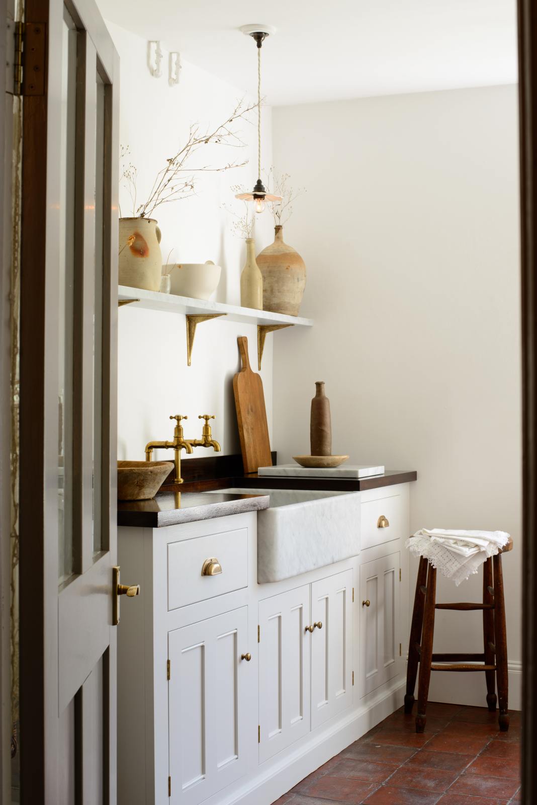An angled view of a simple and functional linen white scullery room with a modest sink run with polished sapele, a large single Carrara marble sink, and aged brass Mayan taps. The warm reds and browns of the terracotta square tiles, vintage furniture, and ceramics serve the room with both a purpose and beautiful aesthetics.