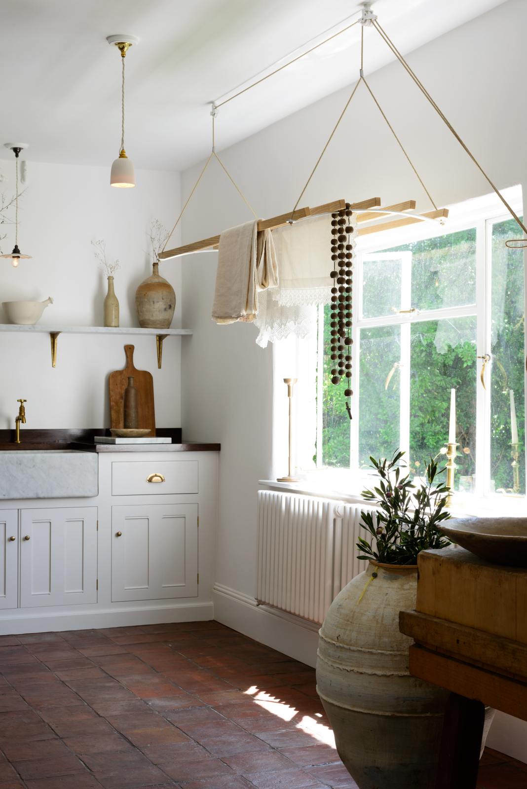 An oblique view of a simple and functional linen white scullery room with a modest sink run with polished sapele, a large single Carrara marble sink, and aged brass Mayan taps. The warm reds and browns of the terracotta square tiles, vintage furniture, ceramics, and a deVOL laundry maid serve the room with both a purpose and beautiful aesthetics.