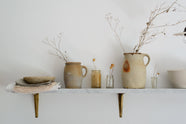 A marble shelf with classic brass brackets holds vintage ceramics, empty jars filled with dried flowers and branches, and muted tablecloths stacked together beneath a textured bowl.
