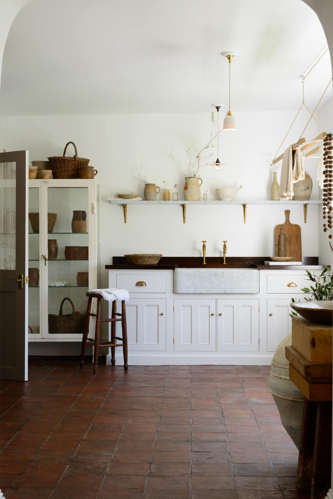 The simple and functional linen white scullery room with a modest sink run with polished sapele, a large single Carrara marble sink, and aged brass Mayan taps. The warm reds and browns of the terracotta square tiles, vintage furniture, ceramics, woven baskets and a deVOL laundry maid serve the room with both a purpose and beautiful aesthetics.