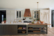 A grand, earth-toned classic kitchen features a Silestone worktop cooker run, glazed wall cupboards, a full-height pantry, a copper canopy, and a large island with a sink, topped with Silestone on one side and copper on the other. Adorned with warm colours such as an aged brass hanging rail, deVOL Heirloom gaselier lights, textured woven baskets, vintage ceramics and loaves of bread, it makes a spectacular use of space but is also down-to-earth.