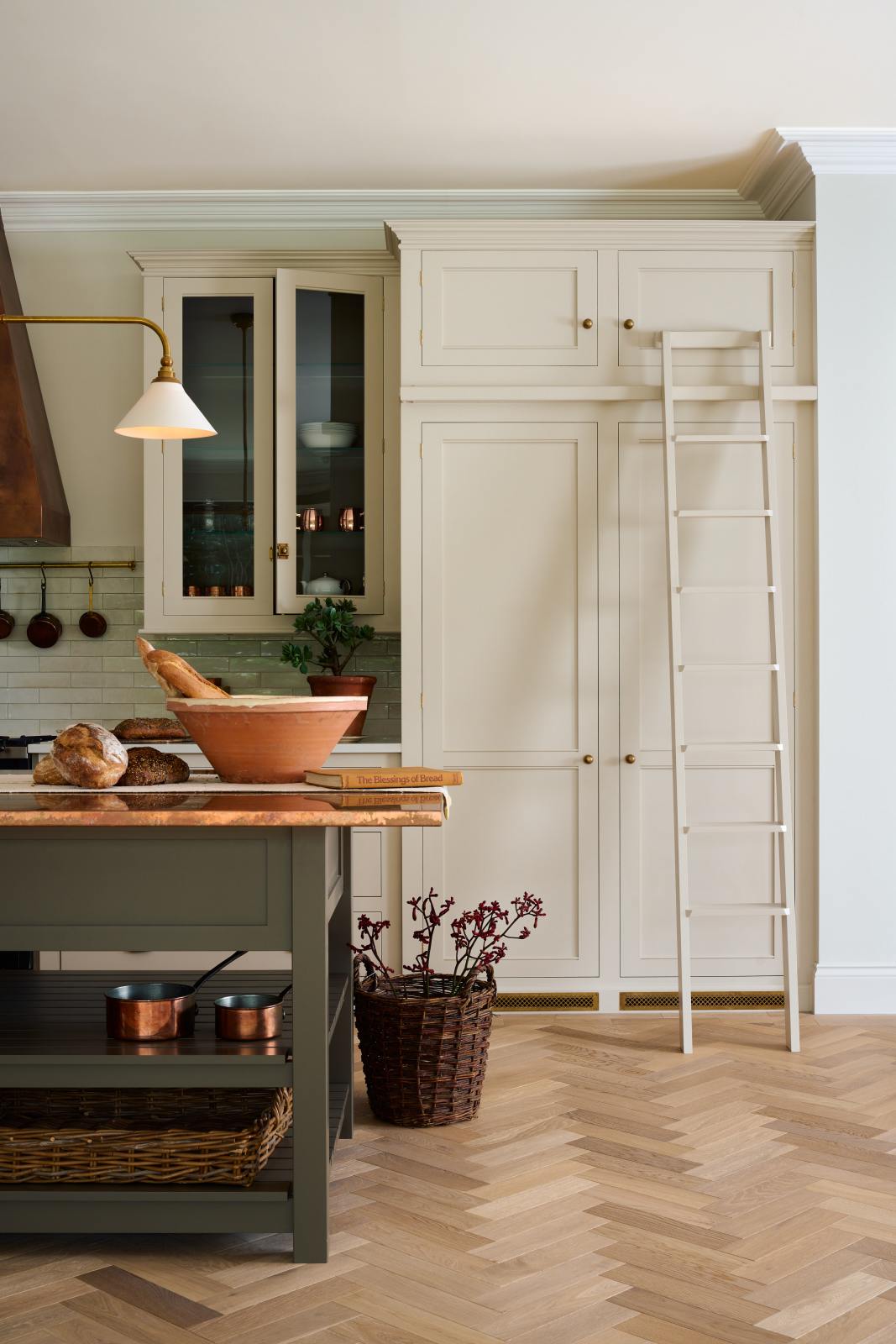 One side of a grand, earth-toned classic kitchen features a Silestone worktop cooker run, glazed wall cupboards, a full-height pantry, a copper canopy, a large island with a copper worktop, and a ladder. Adorned with warm colours such as an aged brass hanging rail, deVOL Heirloom gaselier lights, textured woven baskets, vintage ceramics and loaves of bread, it makes a spectacular use of space but also down-to-earth.