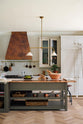 A grand, earth-toned classic kitchen features a Silestone worktop cooker run, glazed wall cupboards, a full-height pantry, a copper canopy, and a large island with a sink, topped with Silestone on one side and copper on the other. Adorned with warm colours such as an aged brass hanging rail, deVOL Heirloom gaselier lights, textured woven baskets, vintage ceramics and loaves of bread, it makes a spectacular use of space but is also down-to-earth.