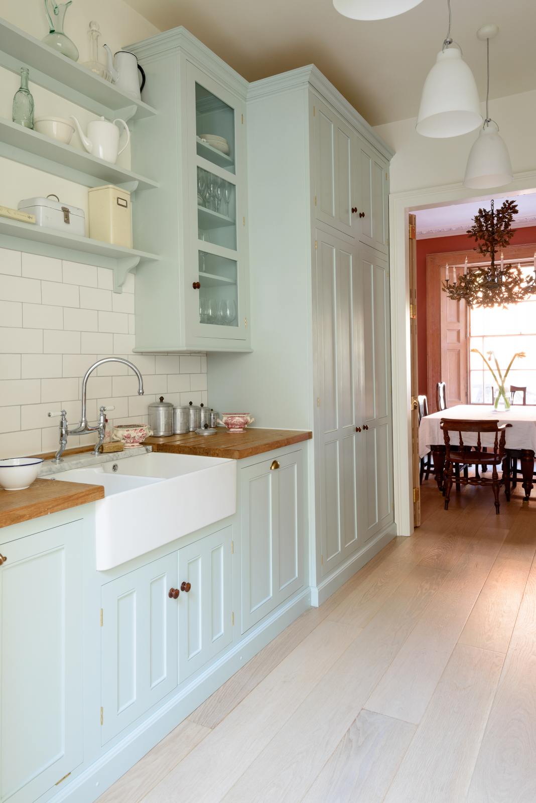 An angled view of a pale-blue classic English galley kitchen features a sink run with a solid oak worktop, a double Belfast sink, a chrome Ionian tap, glazed wall cupboards, pantry cupboards, and shelves. Adorned with artisan ceramics, vintage containers, and simple platters, it creates a humble and practical kitchen that embraces vintage 80s aesthetics.
