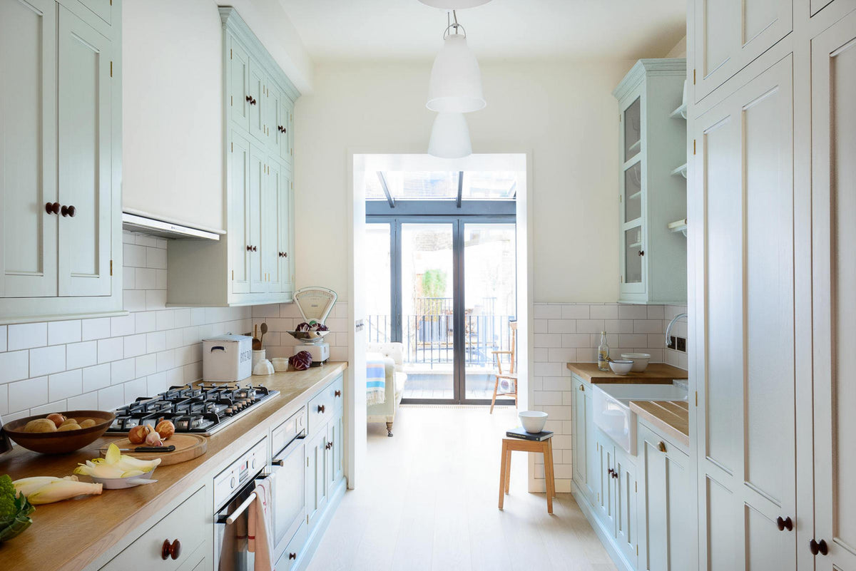 This pale-blue classic English galley kitchen features a cooker and sink run with a solid oak worktop, a double Belfast sink, a chrome Ionian tap, wall cupboards, a canopy, and shelves. Adorned with a vintage scale, a bread tin, artisan ceramics, silver containers, and colourful vegetables, it creates a humble and practical kitchen that embraces vintage 80s aesthetics.