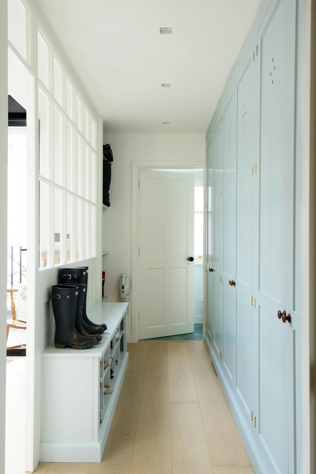 In a bright corridor, the pale-blue coat room features a fitted storage cupboard and shoe shelves. Adorned with muddy boots, a vacuum, and colourful shoes, it creates a nice, tidy, and personalised space.