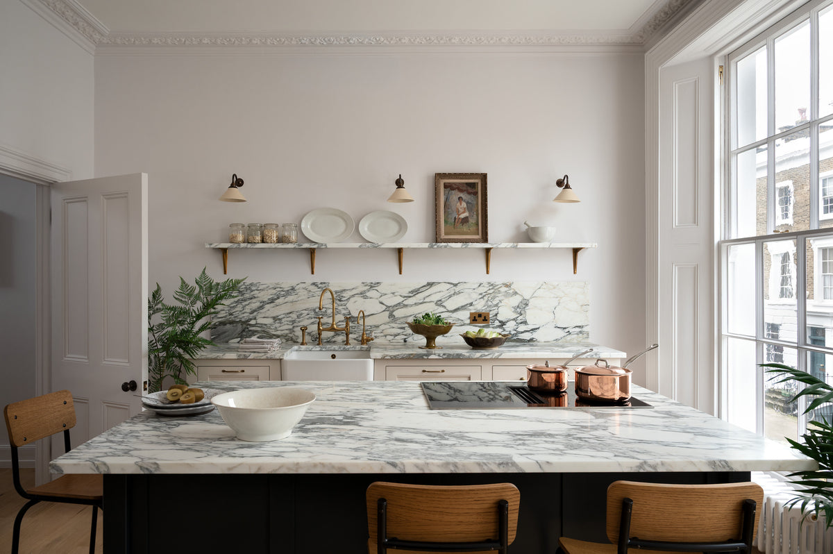 A humble yet elegant off-white Classic English kitchen features a sink run with honed Arabescato marble worktop and splashback, a pantry blue kitchen island with a cooker, mini creamware wall lights, and plumber’s stools. Adorned with artisan ceramics, brass pans, a vintage painting, fresh greens, and grains, creating an intimate social and practical kitchen space.