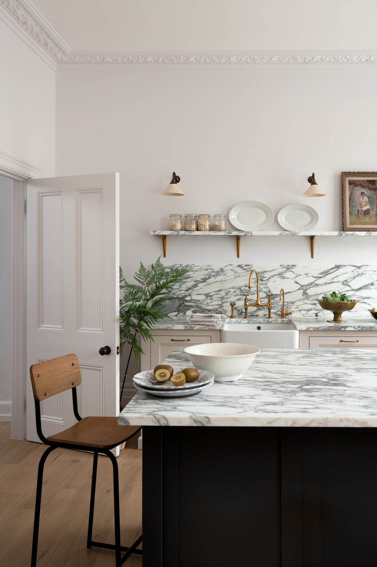 A quiet corner of a humble yet elegant off-white Classic English kitchen features a sink run with honed Arabescato marble worktop and splashback, a pantry blue kitchen island with a cooker, mini creamware wall lights, and a plumber’s stool. Adorned with artisan ceramics, a vintage painting, fresh greens, and grains, creating an intimate social and practical kitchen space.