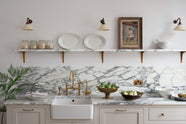 A close-up of a humble yet elegant off-white Classic English kitchen features a sink run with honed Arabescato marble worktop and splashback, a marble shelf, mini creamware wall lights, and an aged brass Ionian tap. Adorned with artisan ceramics, a vintage painting, fresh greens, and grains, creating an intimate social and practical kitchen space.