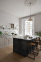 An oblique view of a humble yet elegant off-white Classic English kitchen features a sink run with honed Arabescato marble worktop and splashback, a pantry blue kitchen island with a cooker, mini creamware wall lights, and plumber’s stools. Adorned with artisan ceramics, a vintage painting, fresh greens, and grains, creating an intimate social and practical kitchen space.