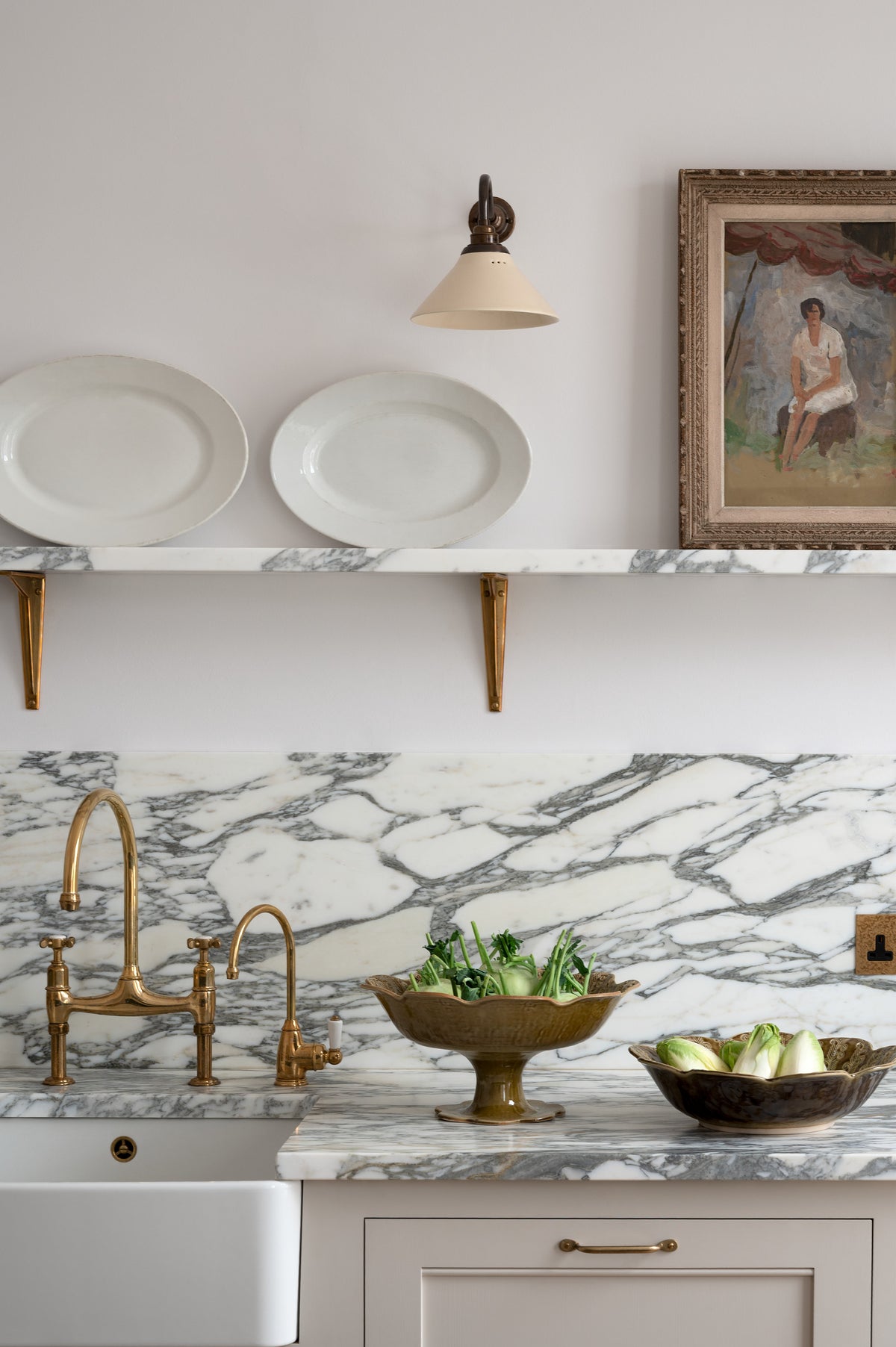 A close-up of a humble yet elegant off-white Classic English kitchen features a sink run with honed Arabescato marble worktop and splashback, a marble shelf, mini creamware wall lights, and an aged brass Ionian tap. Adorned with artisan ceramics, a vintage painting, and fresh greens, creating an intimate social and practical kitchen space.