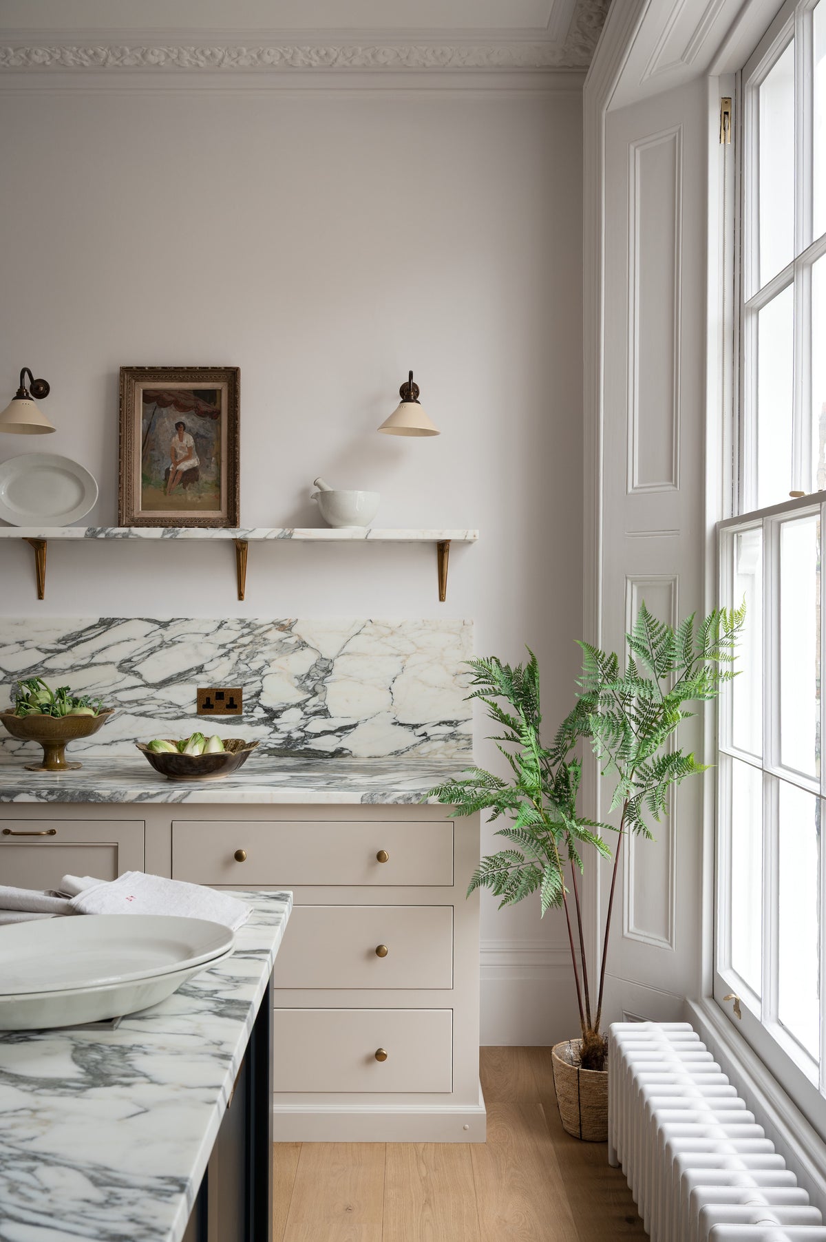 A quiet corner of a humble yet elegant off-white Classic English kitchen features a sink run with honed Arabescato marble worktop and splashback, a pantry blue kitchen island with a cooker, and mini creamware wall lights. Adorned with artisan ceramics, a vintage painting, fresh greens, and grains, creating an intimate social and practical kitchen space.