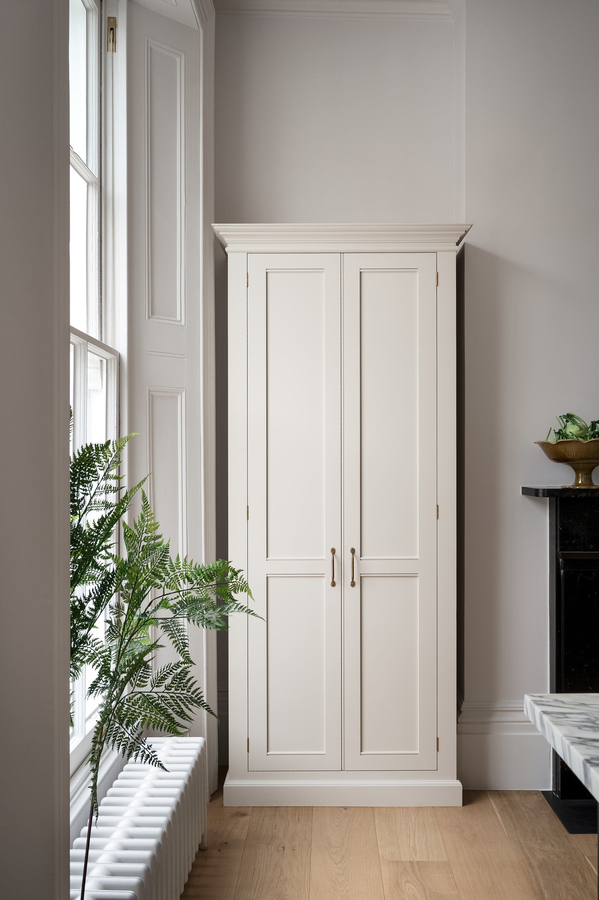 A humble yet elegant off-white Classic English kitchen features a full-height cabinet. Adorned with artisan ceramics, a bowl of fresh artichoke and house plants, creating an intimate social and practical kitchen space.