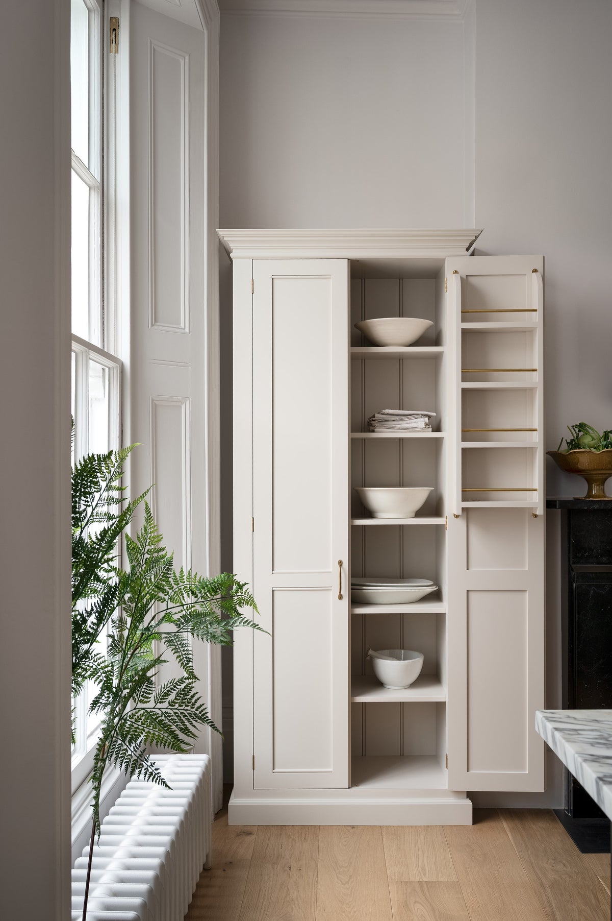 A humble yet elegant off-white Classic English kitchen features a full-height cabinet. Adorned with artisan ceramics, a bowl of fresh artichoke and house plants, creating an intimate social and practical kitchen space.
