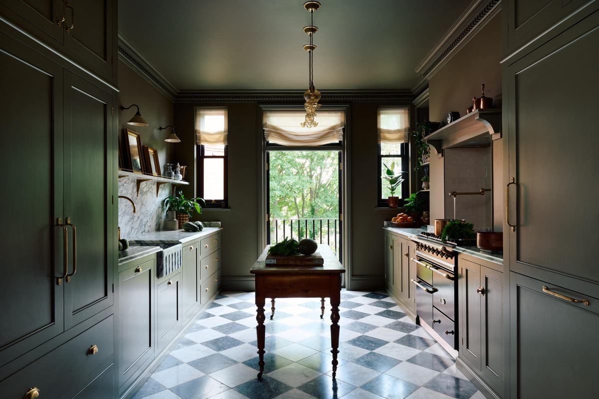 A grand and atmospheric olive green classic English kitchen features a marble worktop and splashback along the sink run, with a single marble sink and an aged brass Ionian tap. A cooker run with an overmantle is paired with an antique prep table on castors. Grand chequerboard marble tiles lead out onto a balcony overlooking green trees.