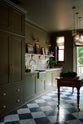 An angled view of a grand and atmospheric olive green classic English kitchen features a marble worktop and splashback along the sink run, with a single marble sink, an aged brass Ionian tap, and an antique prep table on castors. Grand chequerboard marble tiles lead out onto a balcony overlooking green trees.