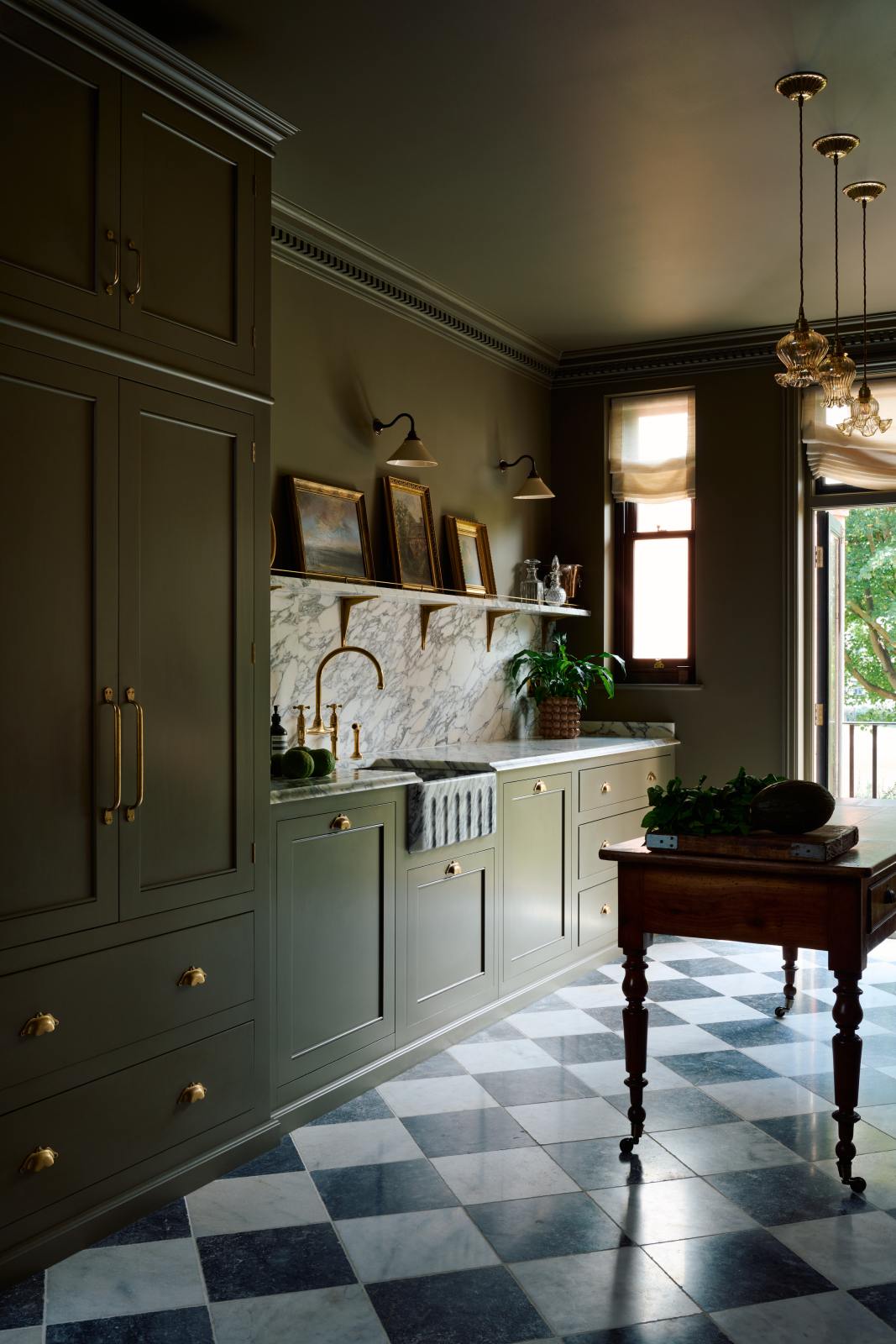An angled view of a grand and atmospheric olive green classic English kitchen features a marble worktop and splashback along the sink run, with a single marble sink, an aged brass Ionian tap, and an antique prep table on castors. Grand chequerboard marble tiles lead out onto a balcony overlooking green trees.