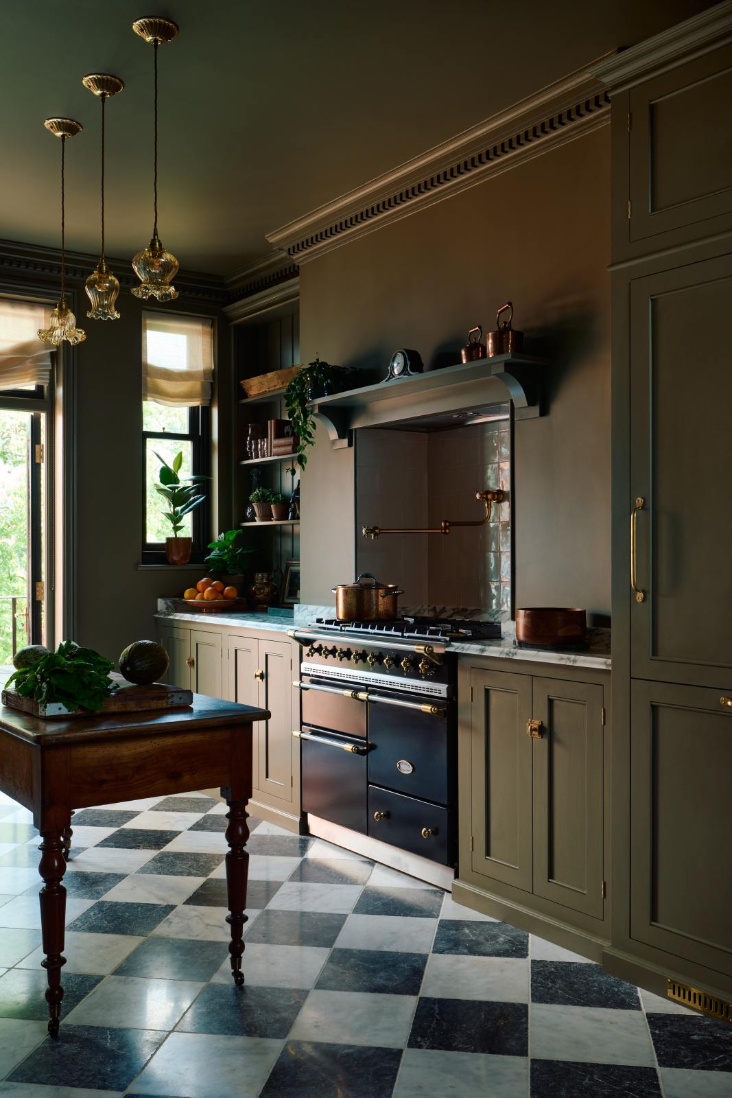 An angled view of a grand and atmospheric olive green classic English kitchen features a cooker run built around the chimney breast, an antique prep table on castors, a full-height fridge/freezer cabinet with top cuboard and an open countertop cupboard filled with beautiful ceramic mugs, a teapot, potted plants, gold-foiled books, glasses, and framed artwork. The black gas cooker in the overmantle has cream subway splashback tiles and a deVOL pot filler tap.