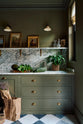 A grand and atmospheric olive green classic English kitchen features a marble worktop, splashback, and shelf along the sink run with a single fluted marble sink, and an aged brass Ionian tap. Two elegant Heirloom wall lights hang above gold-framed paintings, potted plants, vibrant green vegetables, and an assortment of eccentric liquor bottles.