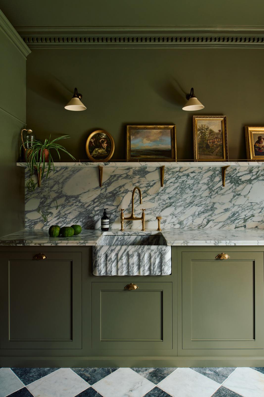 A grand and atmospheric olive green classic English kitchen features a marble worktop, splashback, and shelf along the sink run, a single fluted marble sink and an aged brass Ionian tap. Two beautiful Heirloom wall lights hang above gold-framed famous paintings and potted plants.