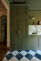 A grand and atmospheric olive green classic English kitchen features a marble worktop, splashback, and shelf along the sink run, a single fluted marble sink and an aged brass Ionian tap. A full-height pantry cabinet takes centre stage, while beside it, beautiful Heirloom wall lights hang above gold-framed paintings and potted plants.