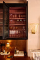 A closer look at a painter’s black classic English café section featuring glazed, reeded cupboards with a blush interior colour and cabinets with aged copper worktop and splashback. A very elegant and sophisticated area in the dining room, complete with a silver coffee machine with a milk frother, as well as wine, cocktail, and scotch glasses.