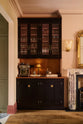 One side of a grand dining room with a painter’s black, classic English café-style nook, reeded glazed cupboards, and aged copper worktops and splashback. A gilded mirror rests above a marble-framed fireplace, framed by amber-frilled sconces. An elegant and sophisticated area in the dining room, complete with a silver coffee machine with a milk frother, as well as wine, cocktail, and scotch glasses.