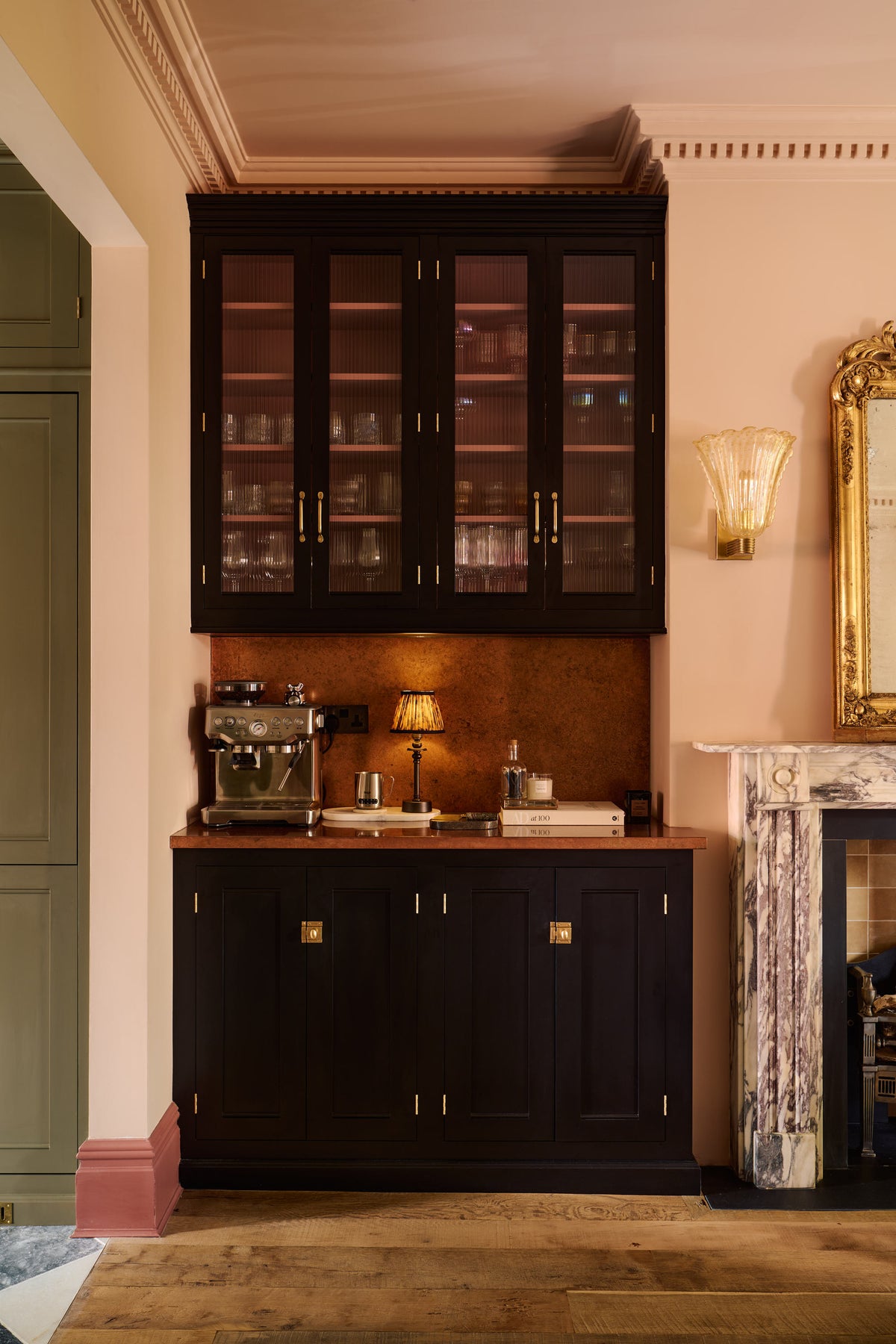 One side of a grand dining room with a painter’s black, classic English café-style nook, reeded glazed cupboards, and aged copper worktops and splashback. A gilded mirror rests above a marble-framed fireplace, framed by amber-frilled sconces. An elegant and sophisticated area in the dining room, complete with a silver coffee machine with a milk frother, as well as wine, cocktail, and scotch glasses.