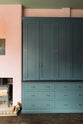 The playful pastel yet classic kitchen features a large Clerkenwell blue full-height pantry cupboard with a black granite worktop and a pink chimney with a small iron wood stove. Decorated with chopped firewood and vintage ceramics, this kitchen has both functionality and personality.
